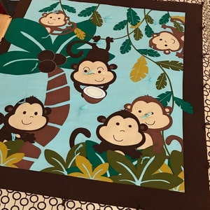 May include: A brown and blue baby blanket with a jungle theme. The blanket features five monkeys, a palm tree, and green leaves.