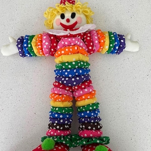 Puede incluir: Mu&ntilde;eca de payaso hecha a mano con sombrero y atuendo de lunares rojos y blancos. Los brazos y las piernas del payaso est&aacute;n hechos de anillos de tela coloridos apilados. El payaso tiene una peluca de hilo amarillo, guantes blancos y zapatos rojos con ribete verde.