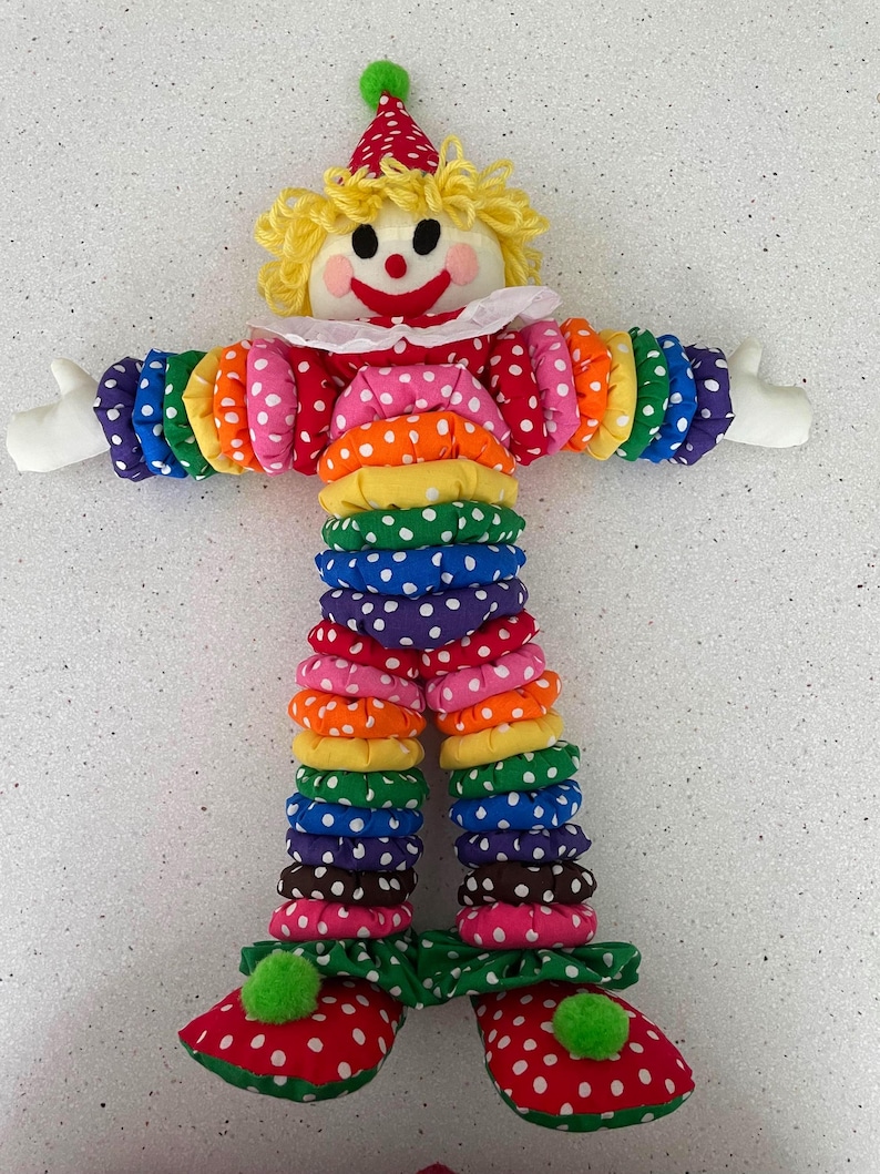 Puede incluir: Mu&ntilde;eca de payaso hecha a mano con sombrero y atuendo de lunares rojos y blancos. Los brazos y las piernas del payaso est&aacute;n hechos de anillos de tela coloridos apilados. El payaso tiene una peluca de hilo amarillo, guantes blancos y zapatos rojos con ribete verde.