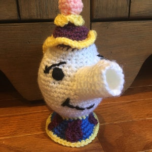 May include: Crocheted teapot character with a white body, purple and yellow hat, and a blue and purple base. The teapot has a long, white spout and black eyes.