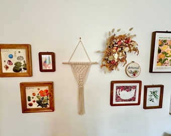 May include: A white wall with a variety of framed artwork and a macrame wall hanging. The artwork includes botanical prints, a cat illustration, and a framed embroidery that says "feminist".