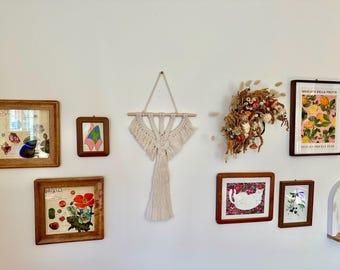 May include: A white wall with a variety of framed artwork, including a macrame wall hanging, a floral print, and a botanical illustration. The wall is decorated with dried flowers and plants.