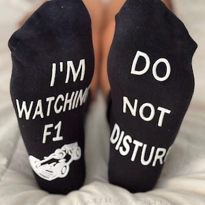 May include: A pair of black socks with white text. One sock says "I'm watching F1" with a small image of a race car. The other sock says "Do not disturb".