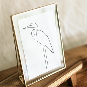 May include: A gold-toned picture frame with a white mat and a line drawing of a bird inside. The frame is sitting on a wooden stand.