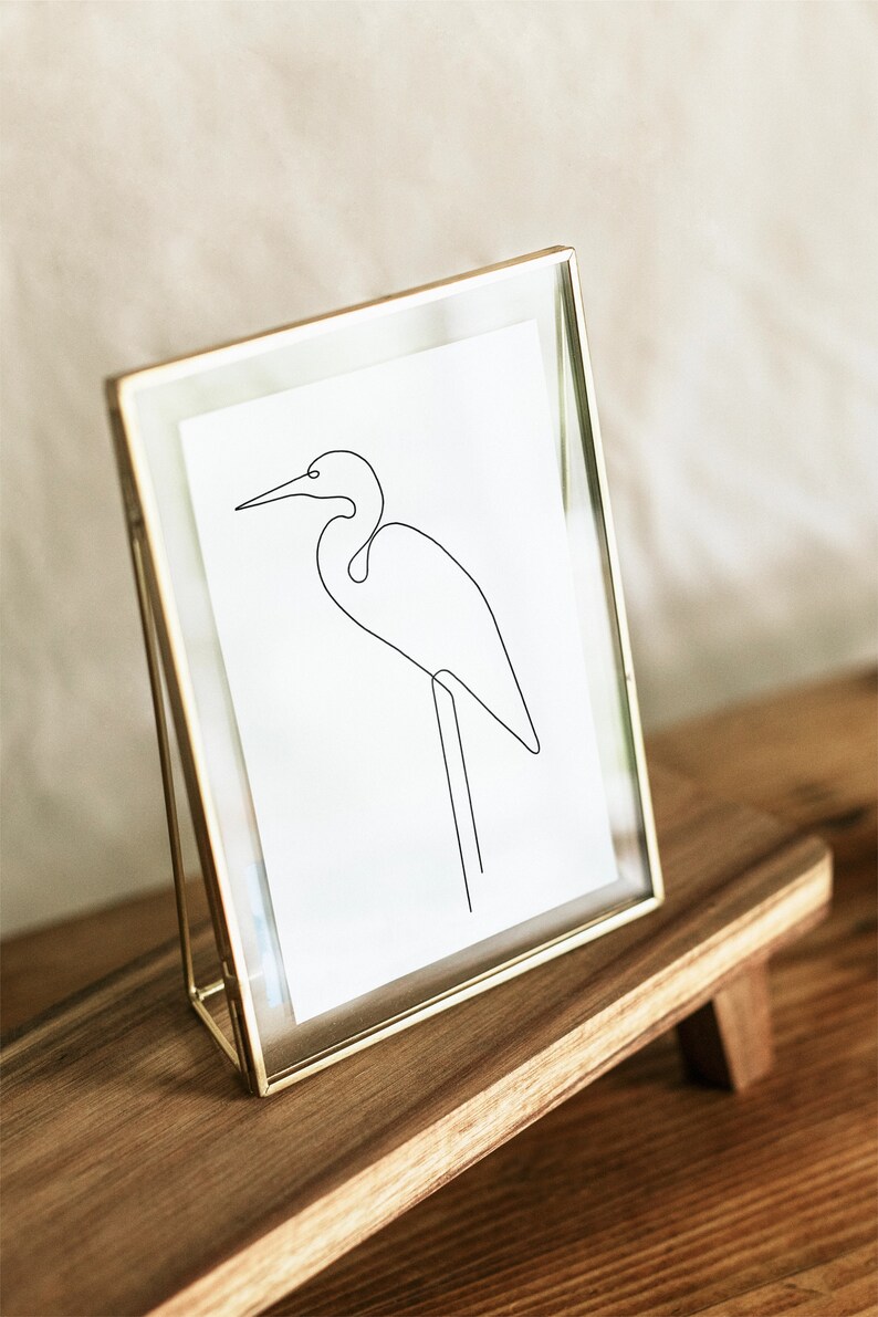 May include: A gold-toned picture frame with a white mat and a line drawing of a bird inside. The frame is sitting on a wooden stand.