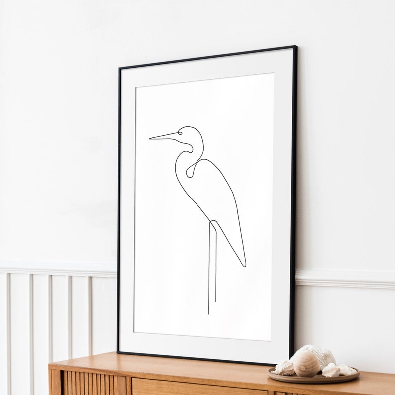 May include: A black framed print of a white egret bird drawn in a single line, standing on one leg. The print is on a white background and is hanging on a white wall.