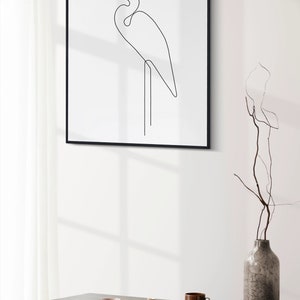 May include: A black framed print of a line drawing of a white egret standing on one leg. The print is hanging on a white wall above a wooden table with a vase, plates, and silverware.