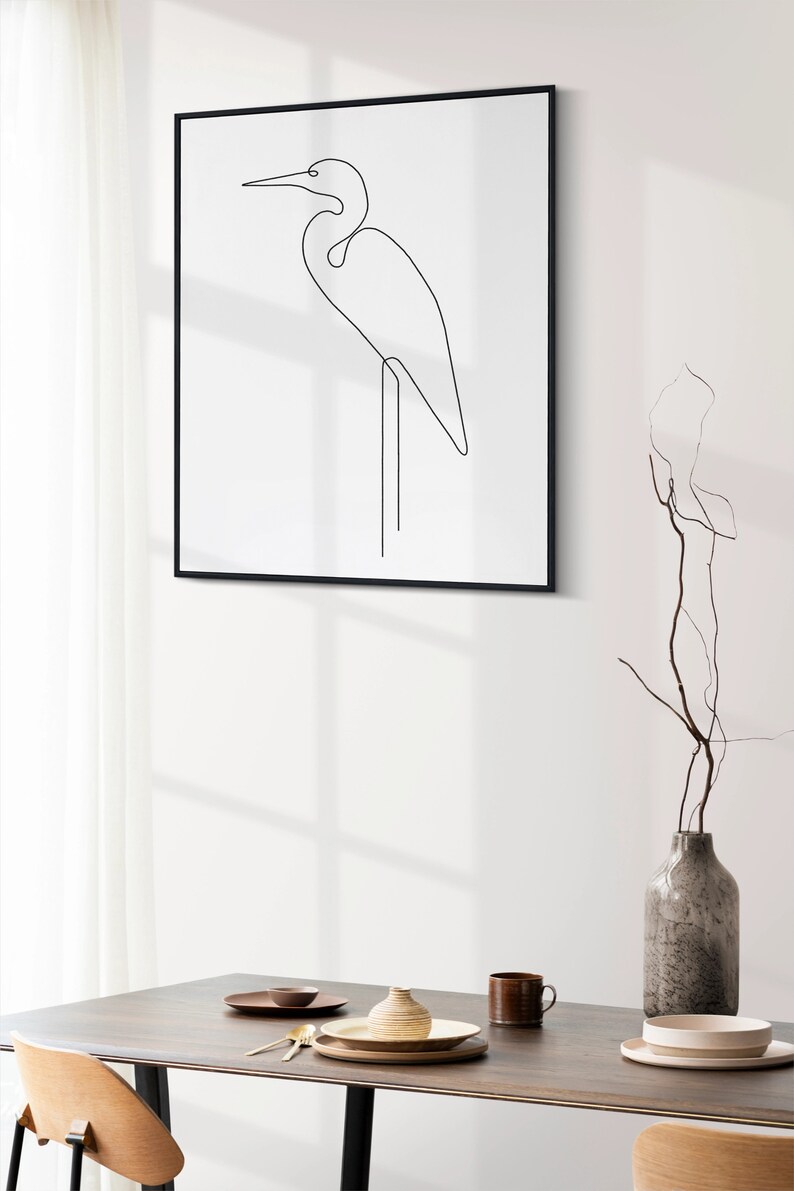 May include: A black framed print of a line drawing of a white egret standing on one leg. The print is hanging on a white wall above a wooden table with a vase, plates, and silverware.