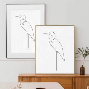 May include: Two minimalist line art prints of a heron or egret bird. The prints are framed in black and gold frames and are hanging on a white wall.