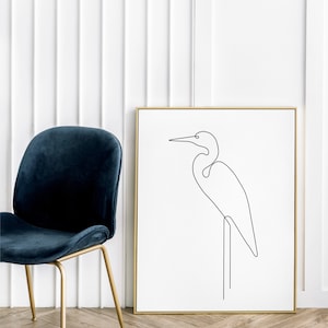 May include: A black and white line drawing of a bird, possibly a heron, standing on a branch. The drawing is framed in a gold frame and is hanging on a white wall.