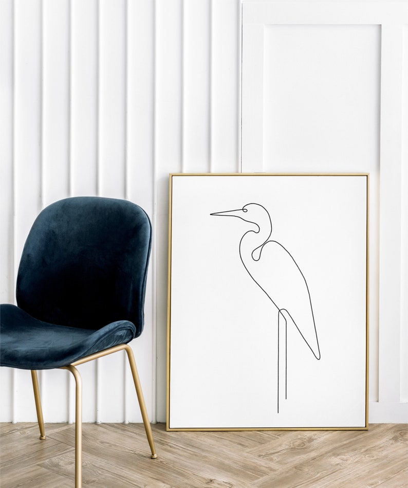 May include: A black and white line drawing of a bird, possibly a heron, standing on a branch. The drawing is framed in a gold frame and is hanging on a white wall.