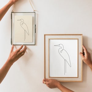 May include: Two framed line drawings of a white egret on a white background. The top frame is black and the bottom frame is light wood.