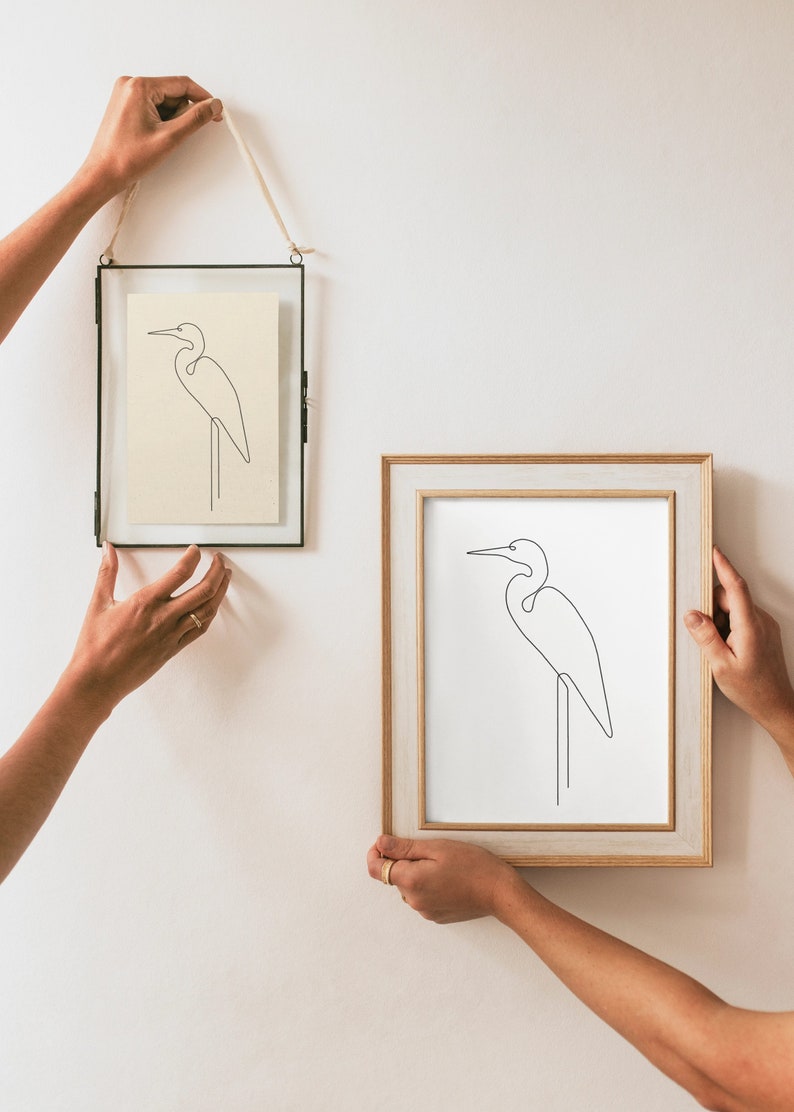 May include: Two framed line drawings of a white egret on a white background. The top frame is black and the bottom frame is light wood.