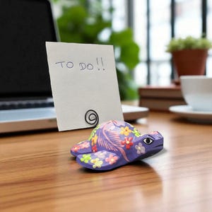 May include: A hand-painted, purple wooden turtle figurine with colorful floral accents. The turtle holds a white note with the text "TO DO!!" and a black paper clip. The figurine is on a wooden desk.