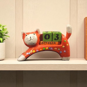 Op de afbeelding: Een decoratieve, oranje katvormige kalender met de datum 3 oktober. De kat heeft een witte kop en staart, met kleurrijke bloemmotieven. De kalenderblokken zijn groen en oranje, wat een speelse toets aan de decoratie geeft.