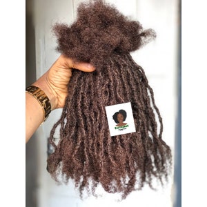 Puede incluir: Un paquete de extensiones de cabello humano marrón en estilo dreadlocks. El cabello es largo y grueso, con una textura suelta y rizada. El cabello se sostiene en una mano, y los extremos de los dreadlocks son visibles.