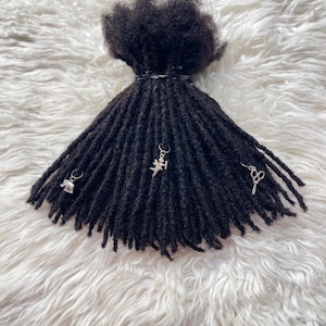 Könnte beinhalten: Ein Bündel schwarzer synthetischer Dreadlocks mit texturierter, gewundener Optik. Die Dreadlocks sind oben mit einem schwarzen Band zusammengebunden und mit drei silberfarbenen Metallanhängern in Form eines Elefanten, eines Engels und einer Schere verziert.