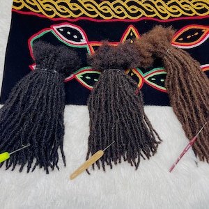 May include: Three bundles of dreadlocks in black, dark brown, and light brown, each with a crochet hook. The dreadlocks are arranged on a white surface with a black textile featuring colorful embroidery in the background.