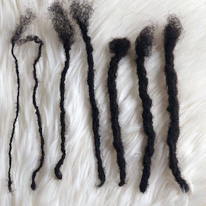 May include: Seven black dreadlocks of varying lengths, displayed on a white fluffy surface.