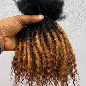 Puede incluir: Un paquete de extensiones de cabello rizado negro y marr&oacute;n. El cabello se mantiene unido en la parte superior, con las ra&iacute;ces de color negro s&oacute;lido. Los rizos hacen la transici&oacute;n a un tono marr&oacute;n m&aacute;s claro hacia los extremos, creando un efecto resaltado. El cabello est&aacute; peinado con rizos apretados y definidos.