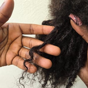 Peut inclure: Gros plan sur une main tenant une section de cheveux bouclés noirs. Les cheveux sont coiffés en un style lâche et texturé.
