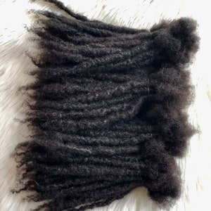 Puede incluir: Primer plano de extensiones de rastas de color marr&oacute;n oscuro. Las extensiones est&aacute;n enrolladas y texturizadas, con un acabado mate natural. Las extensiones de cabello est&aacute;n dispuestas sobre una superficie blanca y esponjosa, creando un contraste de texturas.
