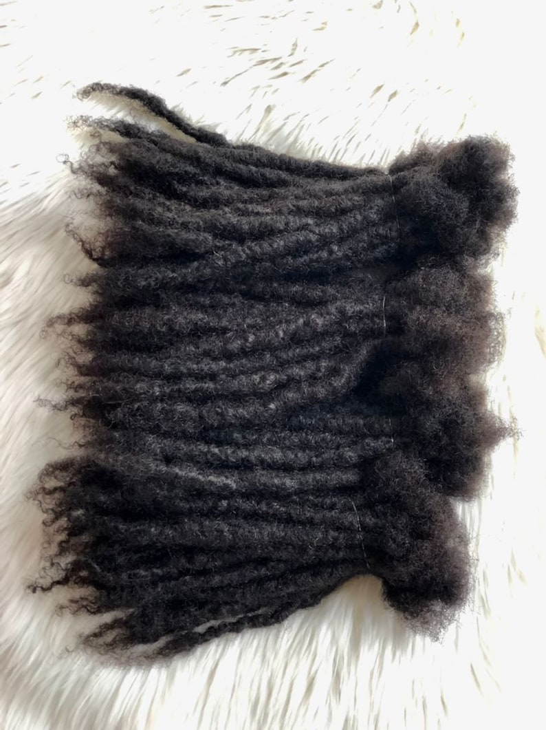 Puede incluir: Primer plano de extensiones de rastas de color marr&oacute;n oscuro. Las extensiones est&aacute;n enrolladas y texturizadas, con un acabado mate natural. Las extensiones de cabello est&aacute;n dispuestas sobre una superficie blanca y esponjosa, creando un contraste de texturas.