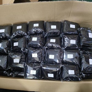 May include: A cardboard box filled with numerous small, sealed plastic bags containing black beads. The bags are neatly arranged in rows.