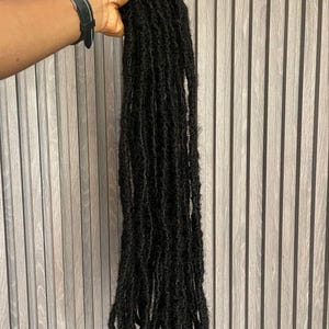 Op de afbeelding: Een hand houdt een bundel lange, zwarte dreadlocks vast. De dreadlocks zijn dik en getextureerd, met een natuurlijk uiterlijk. De achtergrond is een lichtgrijze, verticaal beklede muur. Een zwart horloge is zichtbaar.