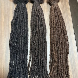 Może przedstawiać: Trzy wiązki dredów w różnych odcieniach brązu i czerni. Dready są ciasno zwinięte i wydają się być wykonane z naturalnych włosów. Wiązki są związane u góry cienką opaską.