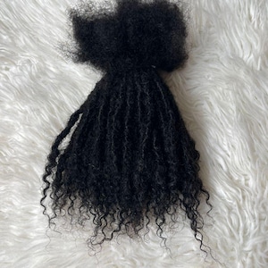 Puede incluir: Un paquete de extensiones de cabello negro, rizado y apretado. El cabello est&aacute; peinado con un patr&oacute;n natural de tipo afro con mechones individuales muy enrollados. El cabello est&aacute; dispuesto sobre una superficie blanca y esponjosa.