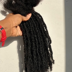 Puede incluir: Un paquete de rastas marrones oscuras y texturizadas. Las rastas se sostienen en una mano, con un reloj rojo visible en la mu&ntilde;eca. El cabello parece natural y sin procesar.