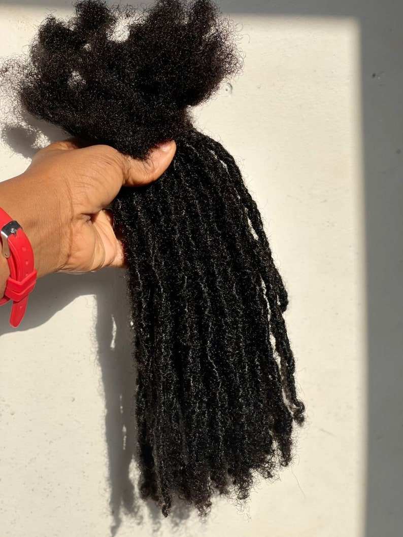 Puede incluir: Un paquete de rastas marrones oscuras y texturizadas. Las rastas se sostienen en una mano, con un reloj rojo visible en la mu&ntilde;eca. El cabello parece natural y sin procesar.