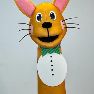 May include: A plush, orange rabbit puppet with pink inner ears, black whiskers, and a black nose. It has a green and white striped bow tie and a white, button-down shirt front. The puppet is designed for hand use.