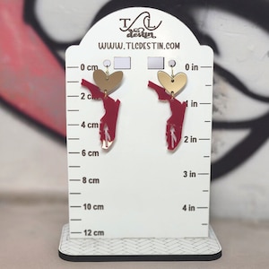 May include: A pair of earrings featuring a gold heart and a red Florida state shape. The earrings are displayed on a white stand with a black and white patterned base. The text "WWW.TLCDESTIN.COM" is visible.