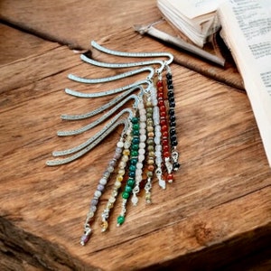 May include: A set of eight silver bookmark holders with beaded tassels. The tassels are made of various semi-precious stones, including amethyst, citrine, green aventurine, rose quartz, carnelian, and black onyx.