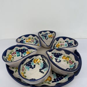 Op de afbeelding: Een witte keramische serveerschaal met een blauwe rand en een bloemenmotief. De schaal heeft zes individuele serveerschalen met blauwe randen en bloemenmotieven. De schalen zijn in een cirkel rond de grotere schaal gerangschikt.