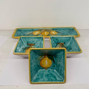 Puede incluir: Un juego de cuatro platos de servir de cerámica con un diseño de limón. Los platos son verdes con limones amarillos y tienen un borde amarillo. El juego incluye un plato de servir rectangular y tres platos cuadrados.