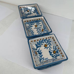 Op de afbeelding: Drie vierkante keramische schalen met een blauwe rand en bloemmotief. De binnenkant van de schalen is wit met blauwe bladeren en oranje bloemaccenten. De schalen zijn op elkaar gestapeld.