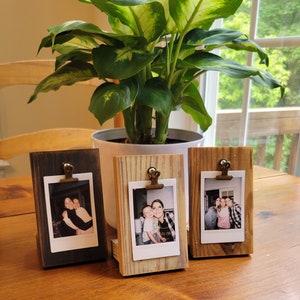 May include: Three wooden picture frames with clips, each holding a Polaroid photo. The frames are in different wood tones, with one being dark brown, one being light brown, and one being a mix of light and dark brown. The photos show people smiling and posing together.