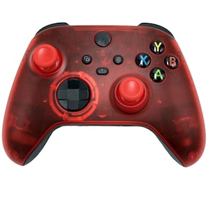 May include: A red and black video game controller with a transparent design. The controller features red thumbsticks, buttons, and a directional pad. The face buttons are colored green, blue, yellow, and red. The controller is designed for gaming.