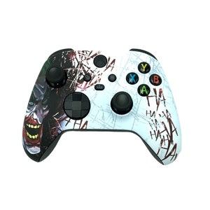 May include: A white Xbox controller with a Joker themed design. The controller features a cracked white background with red and black accents. The Joker's face is on the left side of the controller, and the words "HA HA" are repeated on the right side.