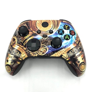 May include: A black and gold Xbox controller with a steampunk design. The controller features a detailed pattern of gears, cogs, and other mechanical elements. The face buttons are colored blue, green, yellow, and red.