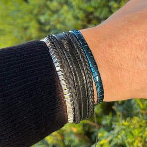 May include: A set of layered bracelets worn on the wrist, featuring a metallic silver-toned studded band, a black textured leather strap, a braided black cord, and a single iridescent blue-green metallic chain-link bracelet with a smooth finish.