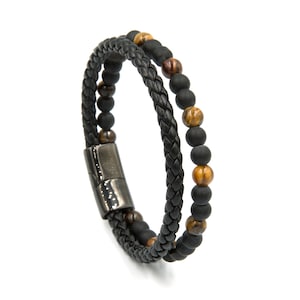 May include: A double-strand bracelet featuring one braided black leather cord and one strand of matte black beads interspersed with brown tiger eye stone beads. The two strands are connected by a polished dark gunmetal magnetic clasp closure.