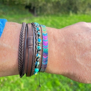 May include: A multi-strand bracelet set featuring a braided brown leather cord, a flat brown leather strap with a central wrap detail, a strand of round turquoise and black marbled beads, and a flat bracelet with a metallic rainbow chevron pattern.