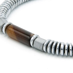 Puede incluir: Una pulsera que presenta una cuenta cilíndrica central de ojo de tigre marrón flanqueada por espaciadores de metal plateado y cuentas grises mate cuadradas, conectadas a una serie de pequeños discos metálicos plateados planos y circulares.