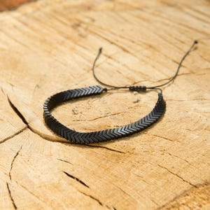 以下が含まれることがあります： 黒いシェブロンデザインのブレスレット。小さな濃い色の長方形のビーズが黒いコードに通されています。ブレスレットは調節可能で、木製の表面に置かれています。