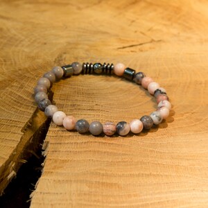 Peut inclure: Un bracelet de perles composé d'une alternance de pierres rondes et polies dans des tons de rose pâle, gris et blanc. Le design intègre une section centrale de petites entretoises cylindriques en métal sombre contrastant avec les perles.