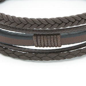 May include: A brown leather bracelet with a braided design. The bracelet has a brown leather strap with a black stripe and a knot in the center.